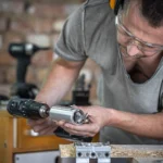 Hombre trabajando en un taller, utilizando un taladro eléctrico para ajustar un accesorio en una pieza de madera, con herramientas y materiales de carpintería visibles en el fondo.